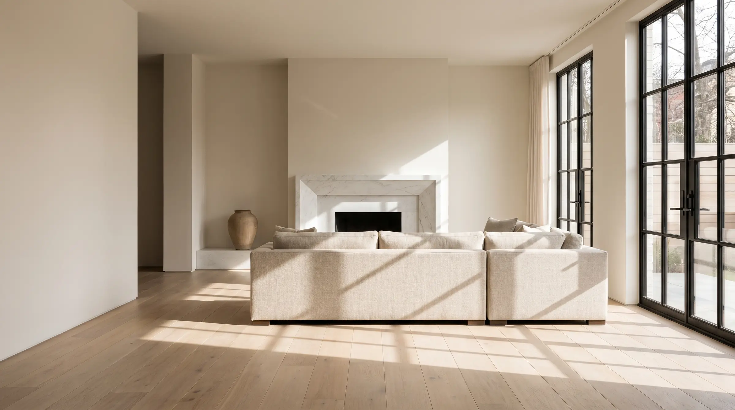 A calm living room with a honed marble fireplace, white oak floors and morning light through steel-framed windows